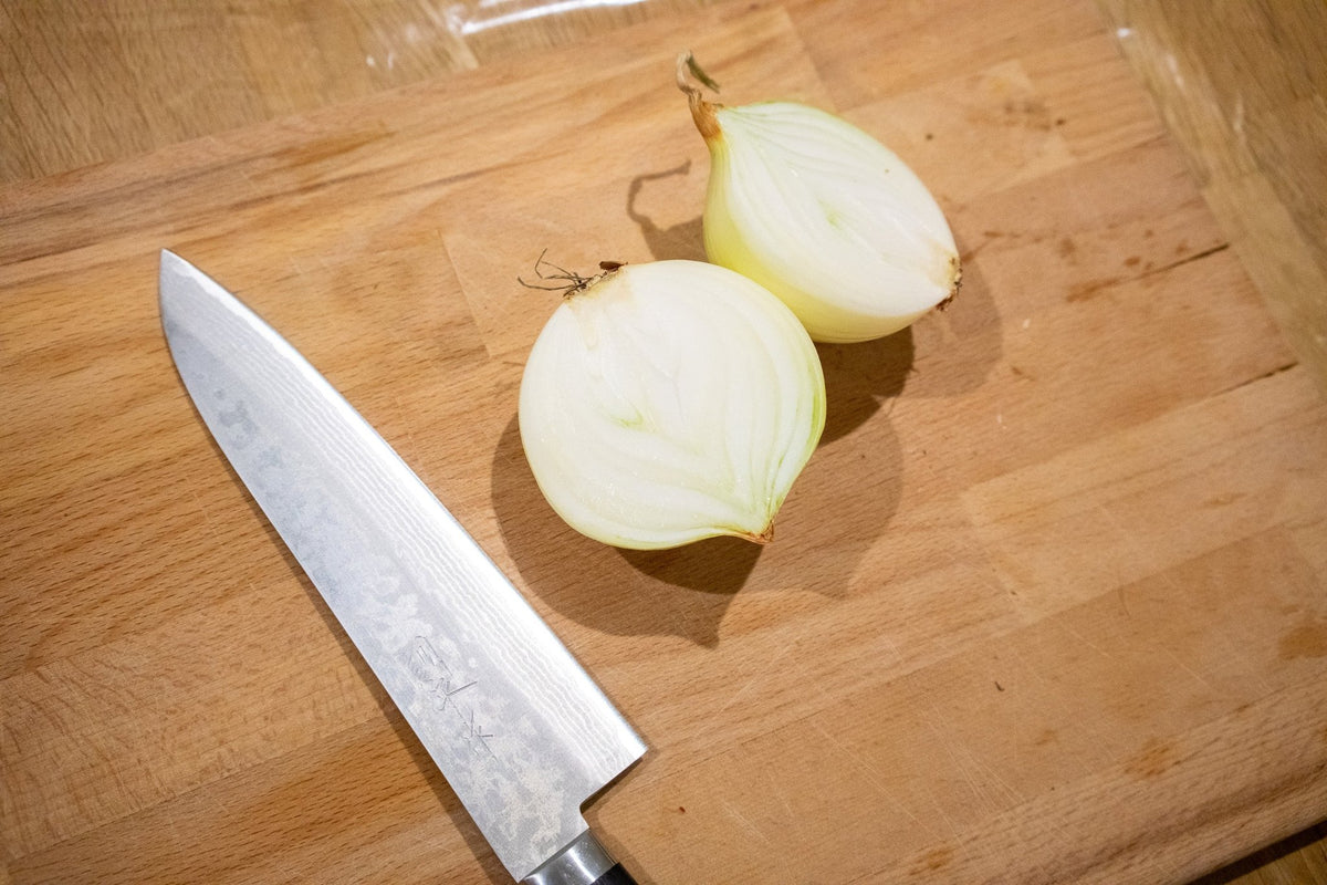 Kokkekniv med damaskusmønster ved siden av en halvert løk på skjærebrett i tre