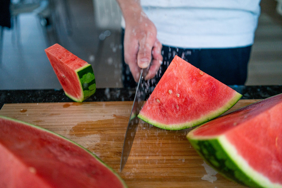Hånd fører en japansk kokkekniv gjennom en vannmelon på et trebrett