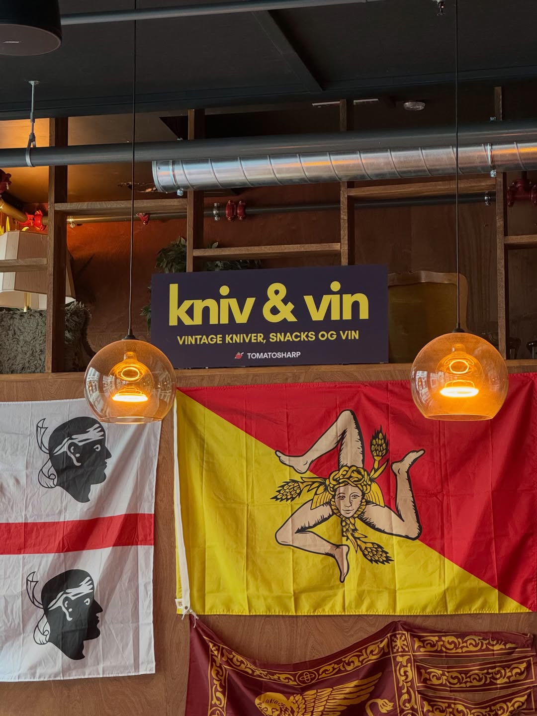 Skilt med Kniv & Vin – vintage kniver, snacks og vin – over flagg og lamper i baren